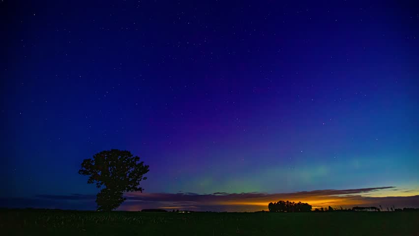 A vast blue starry sky over the fields and trees with an orange sunset on the horizon.