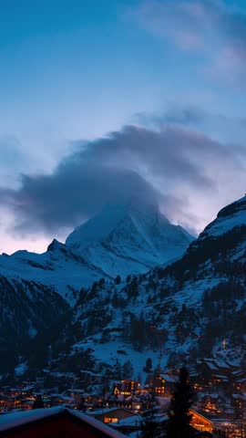 Matterhorn towers over Zermatt, Switzerland, as clouds race by. Snow village lights glow, a hillside cross shines, smoke drifts, and light shifts in a steady vertical frame.