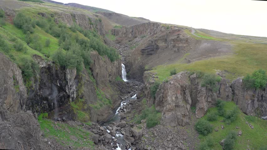 Scenic canyon landscape with small waterfall cascading through rocky cliffs and green hills. A winding stream flows through rugged terrain, surrounded by dramatic volcanic formations under an overcast sky.
