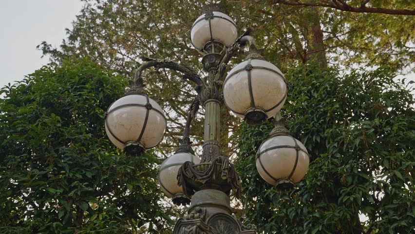 A close-up shot of a classic, ornate street lamp featuring four spherical glass globes, set against lush green trees in a historic Japanese park.