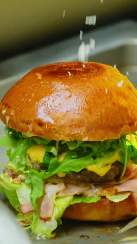 Cook sprinkles freshly grated cheese over assembled burger completing plating. Chef scatters grated cheese across top of burger. Vertical video