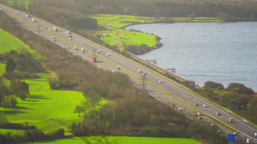 Cars Moving Along M4 Motorway in Wales with Nearby Boating Lake with Lush Green Fields. Illustration of Modern Highway with Natural Surroundings. 4K Clip.