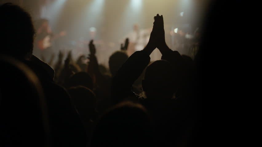 An enthusiastic audience raises hands in silhouette against bright stage lights at a vibrant live music concert or event. The energy builds as hands lift higher, expressing excitement and cheering