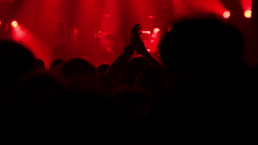 An excited crowd at a live concert, captured from behind the audience. Silhouetted hands are raised and clapping under vibrant red, orange, and yellow stage lights, filmed in slow motion
