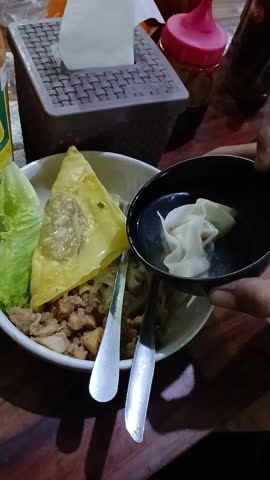 Close-up of a delicious Indonesian street food bowl featuring tofu, dumplings, minced chicken, and fresh lettuce served on a wooden table. A hand holds a small bowl of dumplings, adding an authentic dining moment. Warm lighting enhances textures, creating an appetizing and cozy culinary scene perfect for food-related content.