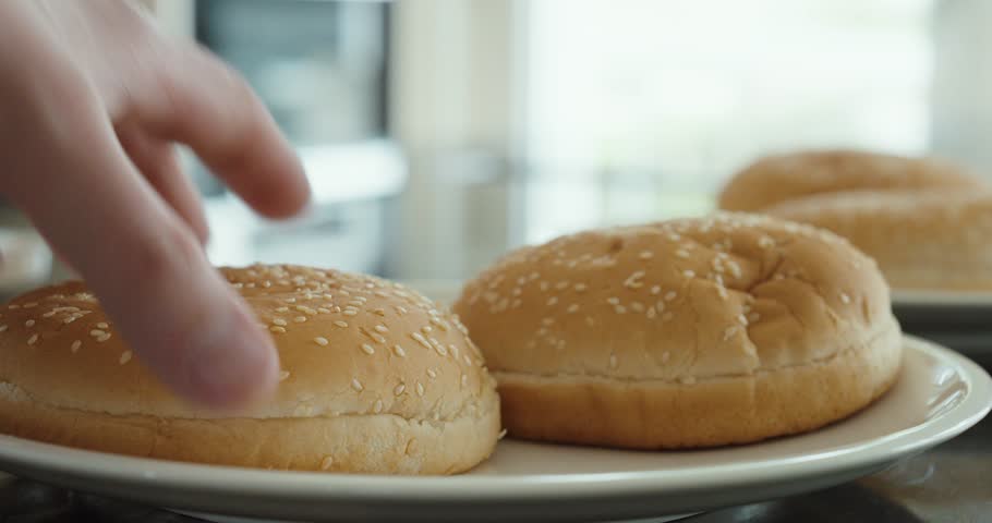 Close-up of a hand picking up a sesame hamburger bun from a plate in a kitchen.