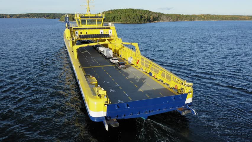 Yellow open deck island car ferry making way ahead in Finnish archipelago. Low altitude aerial view following the vessel at close proximity.