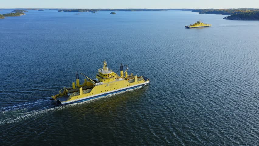 Yellow open deck island car ferry making way ahead in Finnish archipelago. High altitude circling aerial drone view.