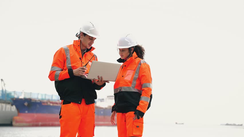 Port workers in safety uniforms using a laptop and radio by a cargo ship. Professional maritime logistics, shipping industry, and international freight transportation management
