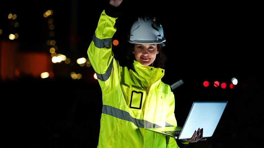 Engineers in hi-vis safety gear collaborate on an industrial project at night, with the woman gesturing while holding a laptop and radio, near a refinery