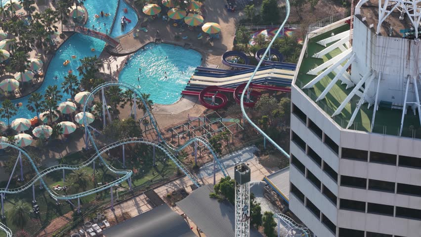Exciting point of view of a person bungee jumping from the rooftop of a tall building over a crowded water park, with roller coasters and pools visible below during the thrilling descent