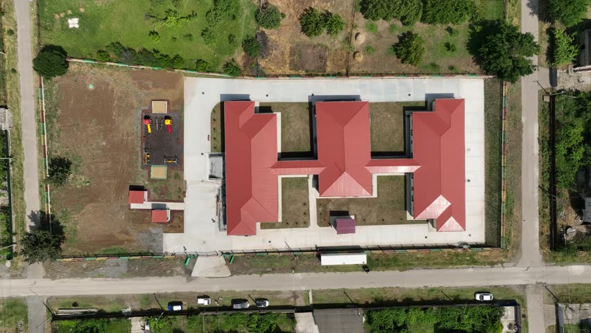 A high-angle drone shot of a newly constructed one-story school or preschool building with a red roof. The facility includes a fenced playground and is situated within a green village community