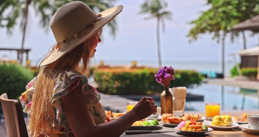 Woman wearing straw hat eating breakfast at luxury tropical resort poolside, ocean view with palm trees and breakfast spread, sunny morning light, luxury travel lifestyle, summer vacation.