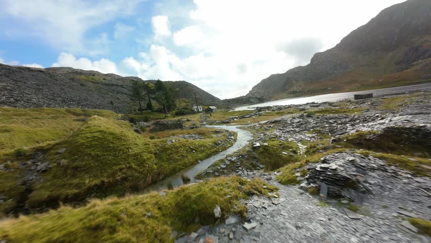 First person view drone flight soaring over a rugged post industrial slate landscape, following a stream into a glistening mountain lake in snowdonia national park, wales