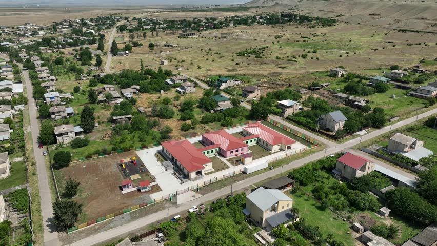 A high-angle drone shot of a newly constructed one-story school or preschool building with a red roof. The facility includes a fenced playground and is situated within a green village community