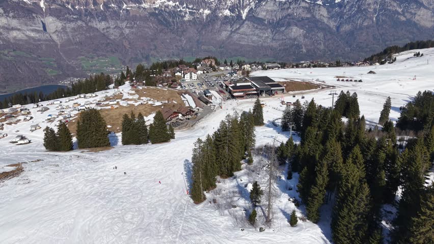 Drone flying toward ski resort base with cars and melting snow. End of winter season atmosphere.