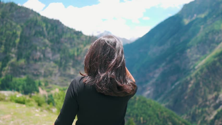 4K shot of back view of girl standing against summer mountains, cold wind blowing her hair. Summer background with copy space. Hiker looking at mountain peak to summit. Adventure, travel concept