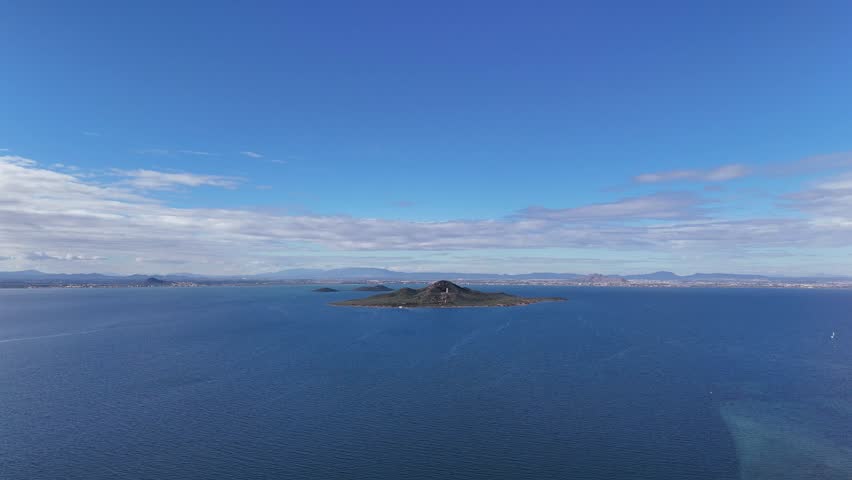 Wide panoramic view of a small island surrounded by calm blue sea with distant coastline and mountains under clear sky. Peaceful nature and travel background.