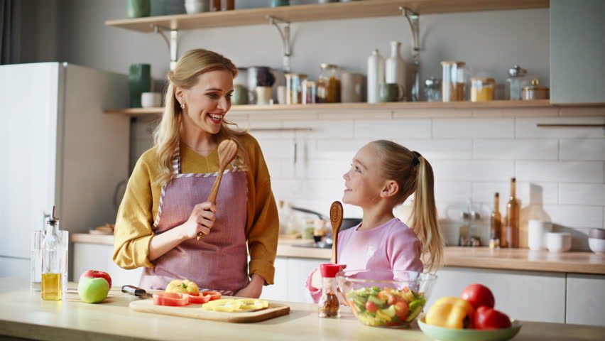 Dancing family having fun cooking dinner in light kitchen feeling joyful closeup. Cheerful woman little girl holding wooden dishes singing together indoors. Overjoyed excited mom child enjoying time 