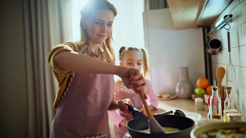 Adorable mom daughter frying pancakes indoor closeup. Caring woman teaching child girl to cook at kitchen. Happy smiling mother kid cooking together at warm light house. Family traditions concept