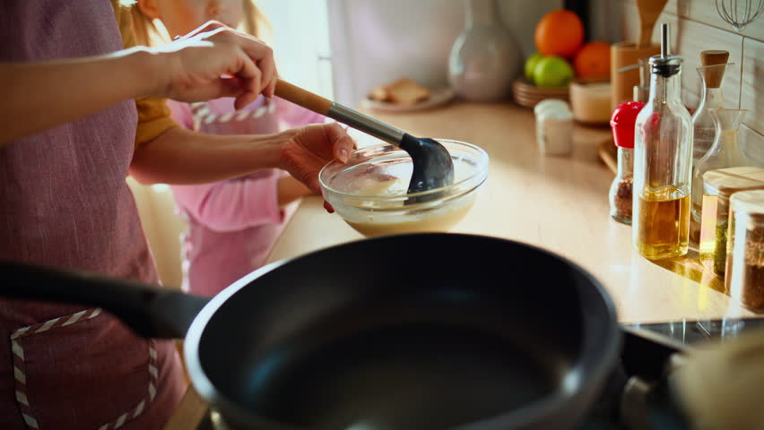 Hand pouring pancakes dough with ladle preparing breakfast at light cuisine room closeup. Blonde family of mom and little child making lunch sunny kitchen. Happy smiling mother teaching small daughter