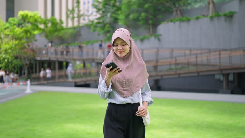 Asian Muslim woman wearing hijab enjoying fresh air outdoors in the city. Attractive young girl standing on balcony, smiling and feeling peaceful, relaxed, and confident in modern urban environment.

