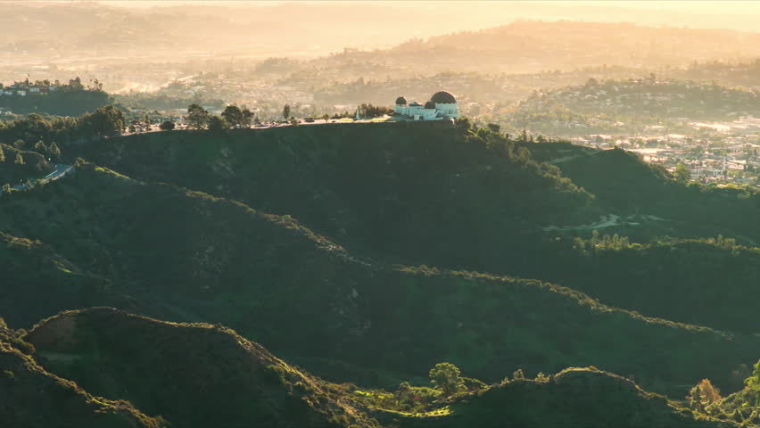 Aerial sunrise view of Griffith Observatory rising above green Hollywood Hills with warm golden haze covering the Los Angeles cityscape and distant skyline.
