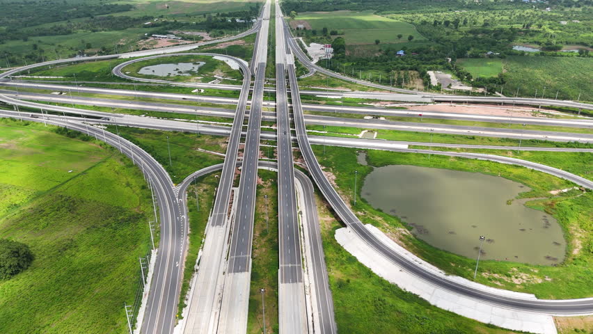 Aerial view of modern highway interchange with multilayer roads and curved ramps surrounded by green farmland, showcasing transportation infrastructure, urban development, and road network planning.