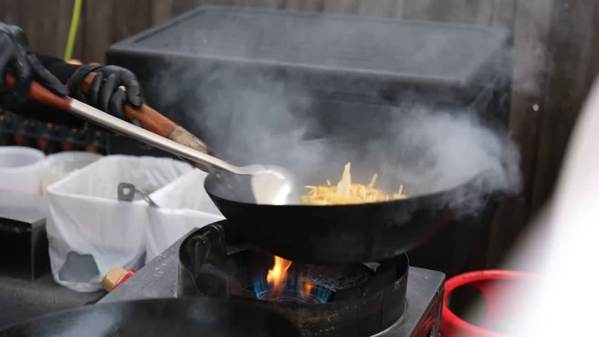 Chef tossing noodles in a wok over a visible blue and orange gas flame. The chef splashes dark soy sauce onto the dish while flipping the food over the intense fire.