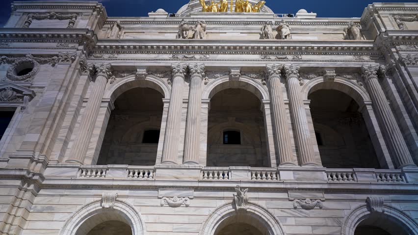 Visitors view the Minnesota state Capitol with bright sunlight highlighting the structure and design in Saint Paul on a clear day