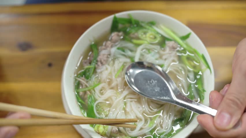 Taster Enjoys Flavorful Beef Broth. Caucasian Critic Tastes Vietnamese Pho With Greens And Tender Beef. An Intimate Shot Capturing Critic Savoring Vietnamese Pho With Greens And Aromatic Broth