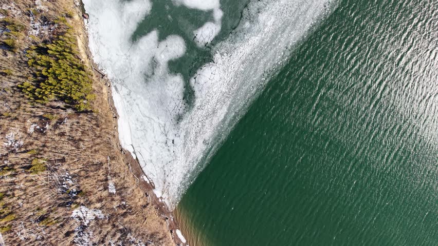 Top down aerial drone view of a lake or river meeting icy shoreline with turquoise green water flowing alongside snow and rocky terrain near Whitefish, Montana.