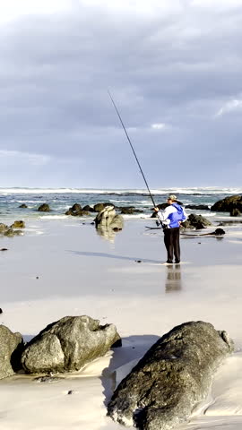 Fisherman stands on beach with rod in hand waiting for fish to bite. Vertical