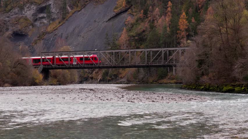 A majestic and iconic cinematic shot of a vibrant red passenger train crossing a high stone bridge or viaduct over a rushing river in the Swiss Alps, surrounded by dense autumn forests and steep mountain cliffs, perfectly capturing the spirit of luxury mountain travel and the incredible engineering feat of high-altitude rail transport in Europe, Red Train Crossing Bridge in Swiss Alps.