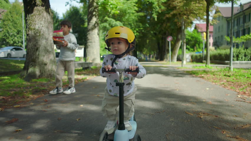 Closeup toddler riding scooter in park wearing yellow helmet, joyful playful moment, older sibling follows behind holding toy airplane, carefree childhood scene