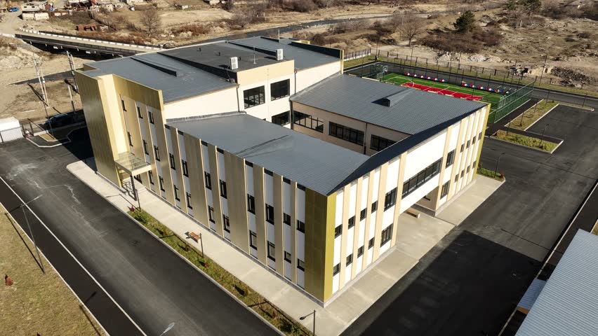 Aerial shot of a contemporary school facility featuring a red and green outdoor sports court, asphalt paths, and surrounding countryside landscape during daytime.