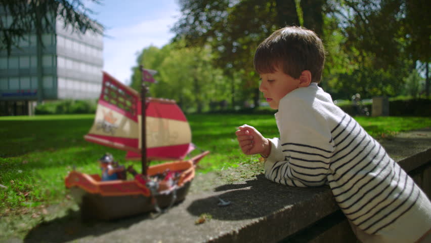 Smiling boy in striped sweater leaning on stone ledge outdoors, looking with joy at toy pirate ship with red sail while enjoying playful moment in park