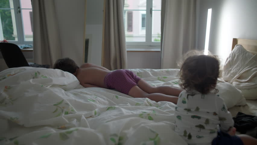 Two siblings playing together on unmade bed, joyful playful moment, childhood bonding, family lifestyle, carefree morning, energetic toddler kicking pillow, happy brotherhood at home
