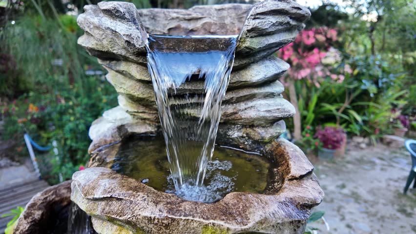 Zoom out decorative stone water fountain cascading in a lush green garden, evoking a peaceful and zen atmosphere.