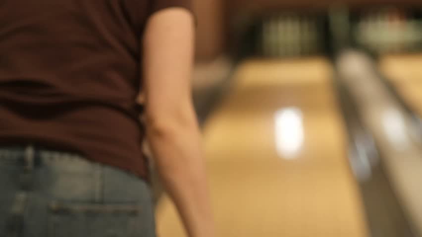 Woman in casual clothing positions herself to bowl with a pink ball in a bowling alley, showcasing the lane and bowling pins in the background