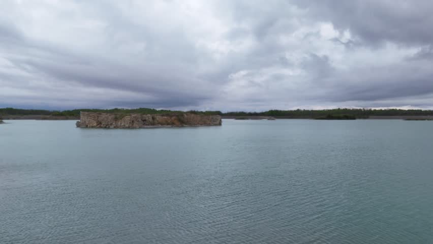 Drone flying low across clear blue water in a limestone quarry in Steep Rock Manitoba toward a limestone island with cliffs visible under cloudy summer skies.