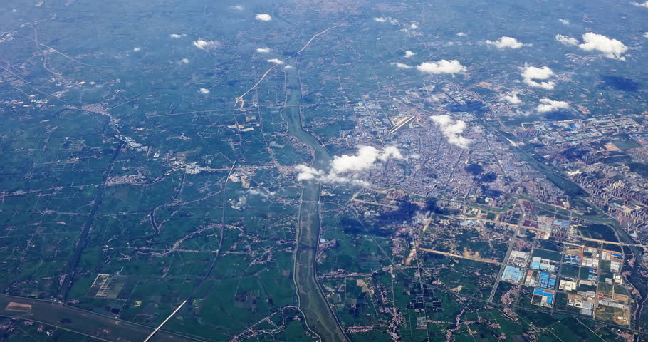Aerial view of a long river winding through a sprawling urban landscape and rural farm fields under a light cloud cover.
