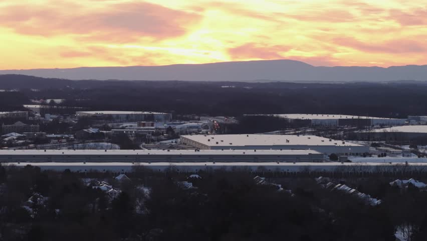 Aerial sunset view over snowy Carlisle, Pennsylvania showing warehouse buildings, shopping center areas and suburban neighborhoods with mountains in distance. Wide shot.