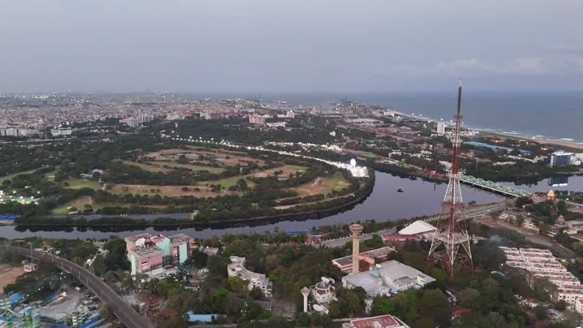 Aerial perspective of central Chennai at dusk, showing the Cooum River flowing toward the sea with Napier Bridge, surrounding roads, and coastal landmarks illuminated by evening lights.