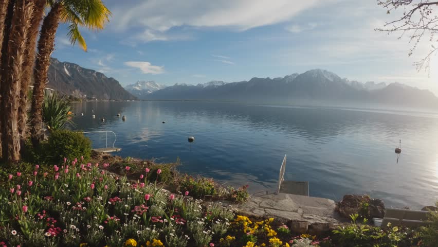 Scenic view of Lake Geneva in Montreux, Switzerland, featuring palm trees and colorful flowers in the foreground, calm reflective water and alpine mountains in the background. Soft sunlight creates a peaceful and premium travel atmosphere.