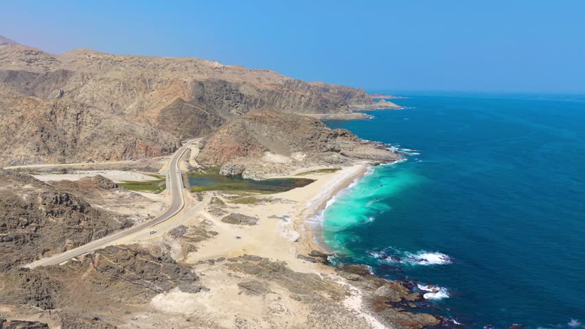 Aerial view of a coastal highway winding between rocky mountains and a sandy beach with turquoise water in Saudi Arabia.