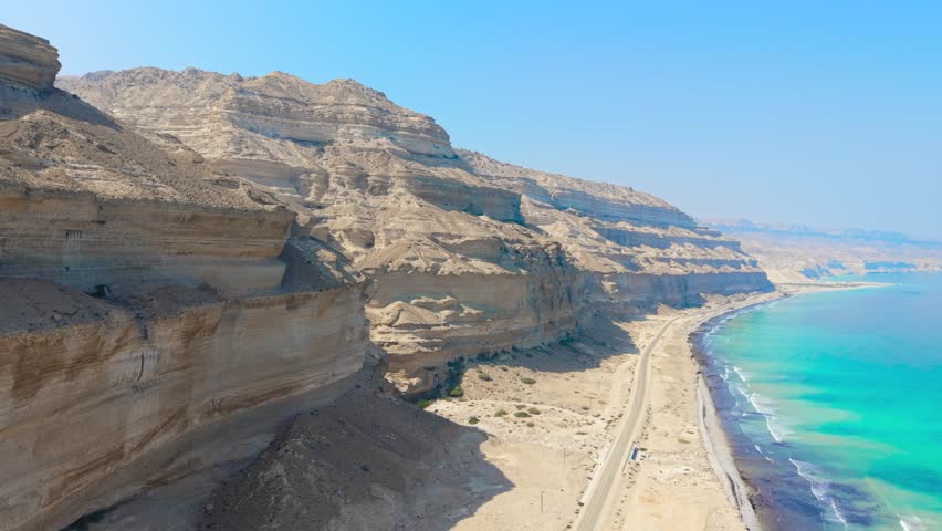 Aerial view of a long asphalt road stretching between massive desert cliffs and the turquoise waters of the Arabian Sea in Salalah, Oman.