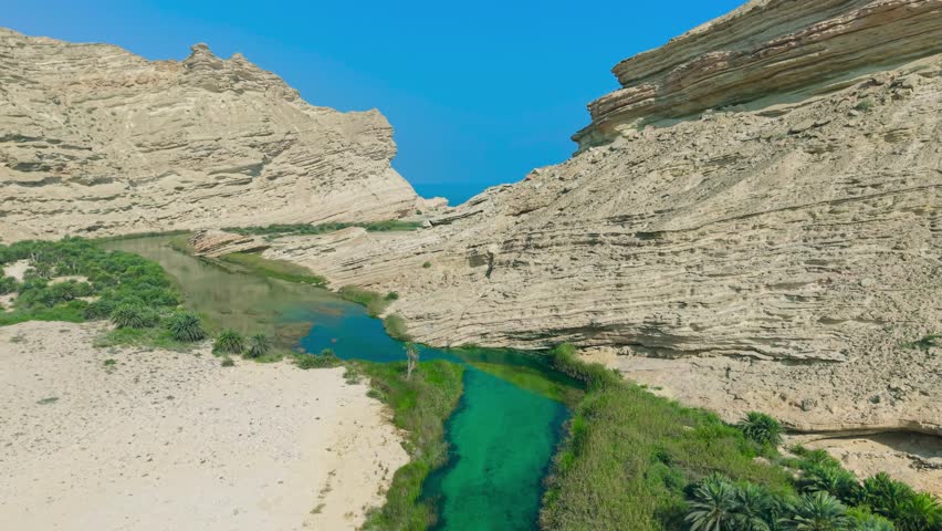 Aerial view of a turquoise river flowing through a desert valley with green palm trees and steep rock cliffs in Wadi Shuwaymiyah, Oman.