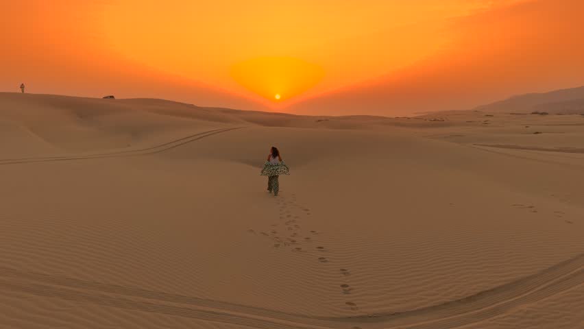 Aerial view of a young woman walking across sand dunes toward the setting sun in the desert of Oman.