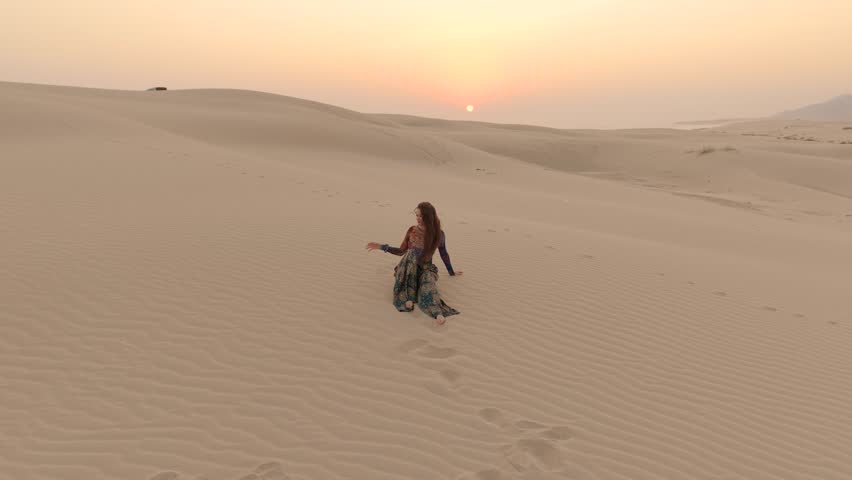 Young woman sitting on ripples of a sand dune during sunset seen from an aerial view in the Sharqiya Sands desert of Oman.